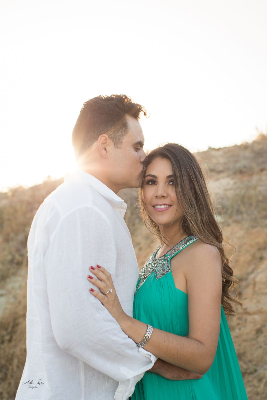 Romantic couple embracing at golden sunset, man kissing womans forehead, woman in turquoise dress.