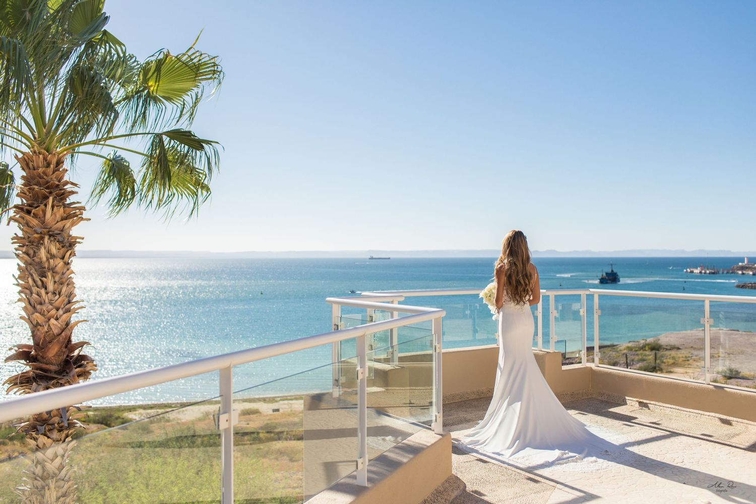Bride in elegant white gown stands on balcony overlooking stunning turquoise ocean waters.
