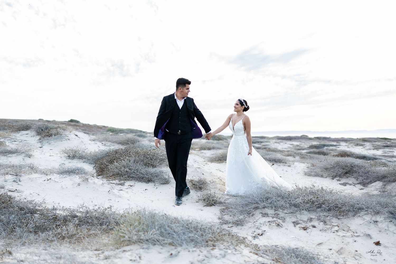 Bride and groom walking hand in hand across white sand dunes at twilight.