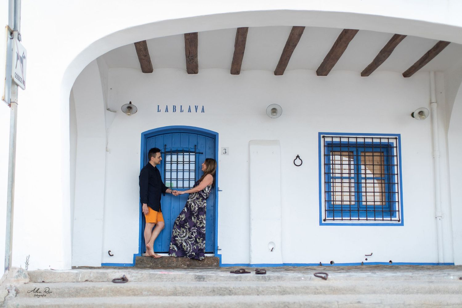 Couple sharing a romantic moment at a white Mediterranean building with blue door and windows.