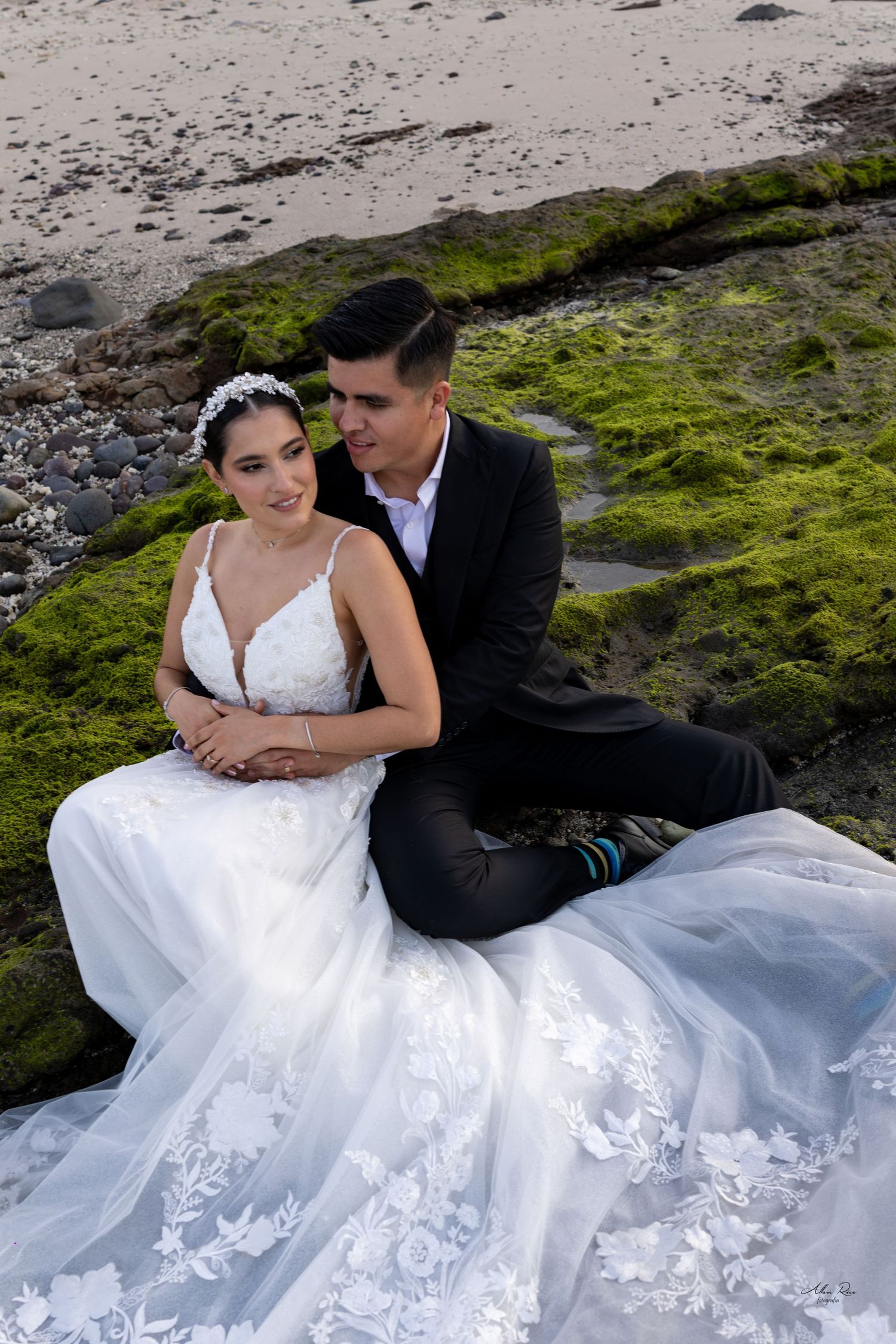 Bride and groom sitting on moss-covered rocks at the beach in wedding attire.