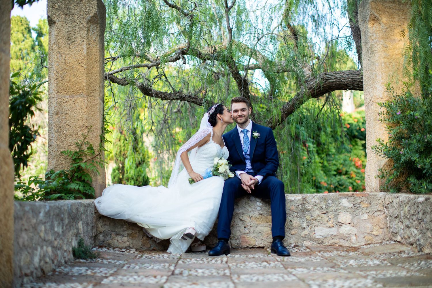Bride and groom sitting on stone bench under scenic trees in elegant wedding attire.