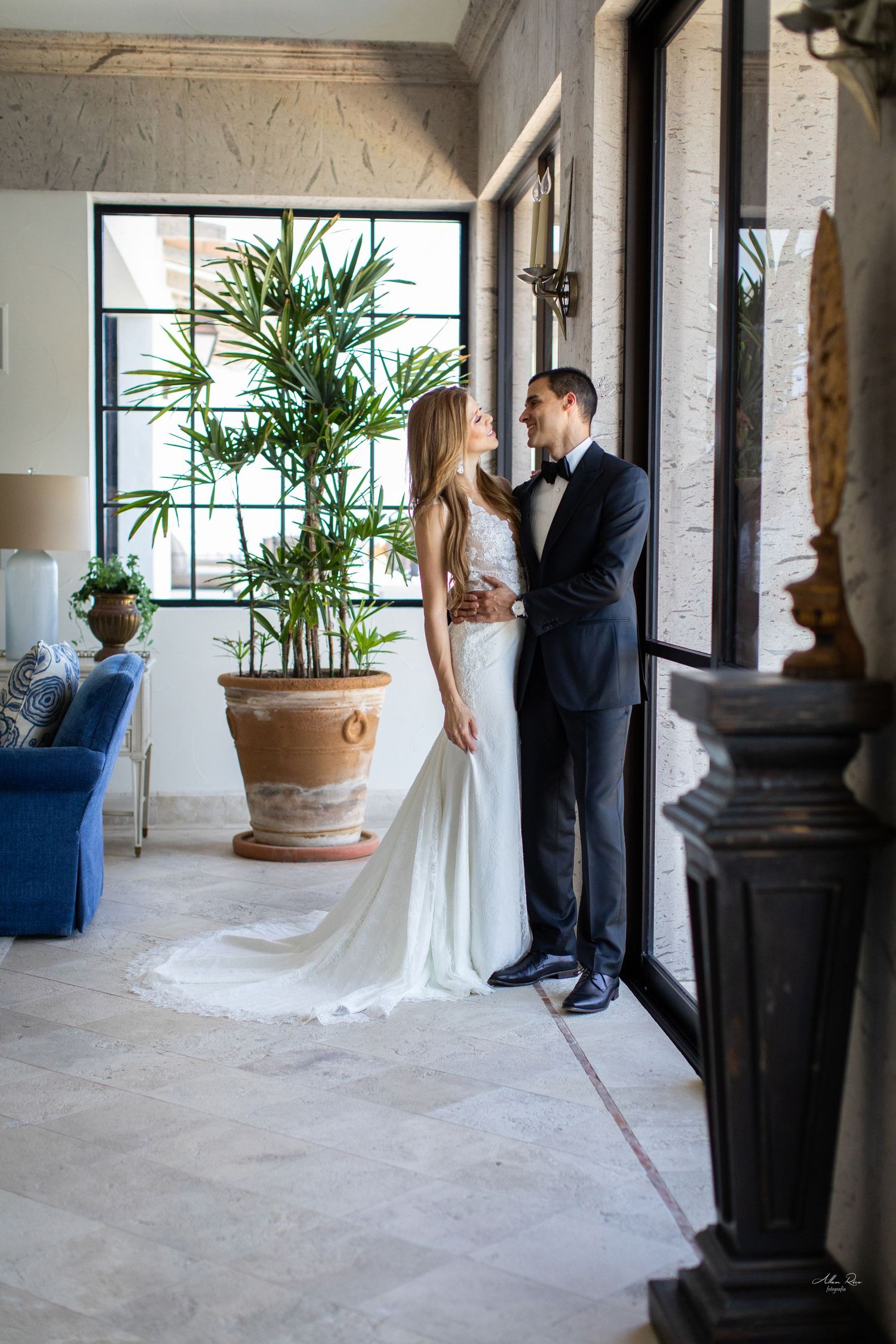 Bride and groom share an intimate moment in elegant interior with large potted palm and blue velvet sofa.