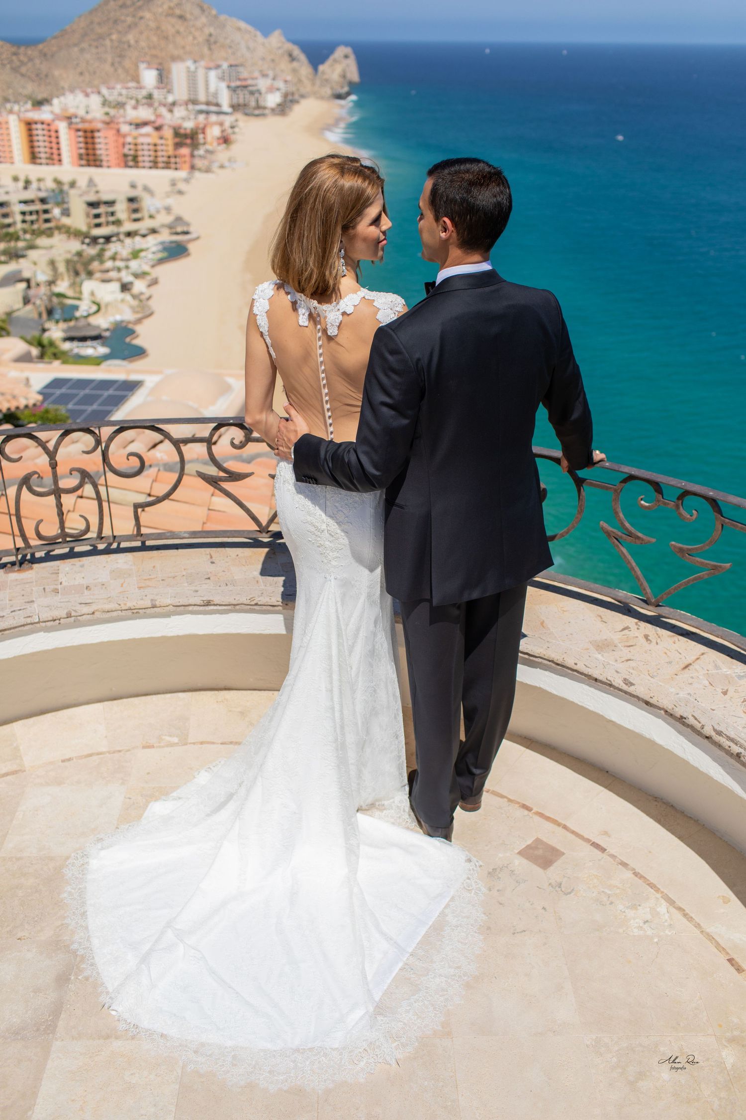 Bride and groom embrace on circular balcony with stunning aerial view of Cabo San Lucas beach and ocean.