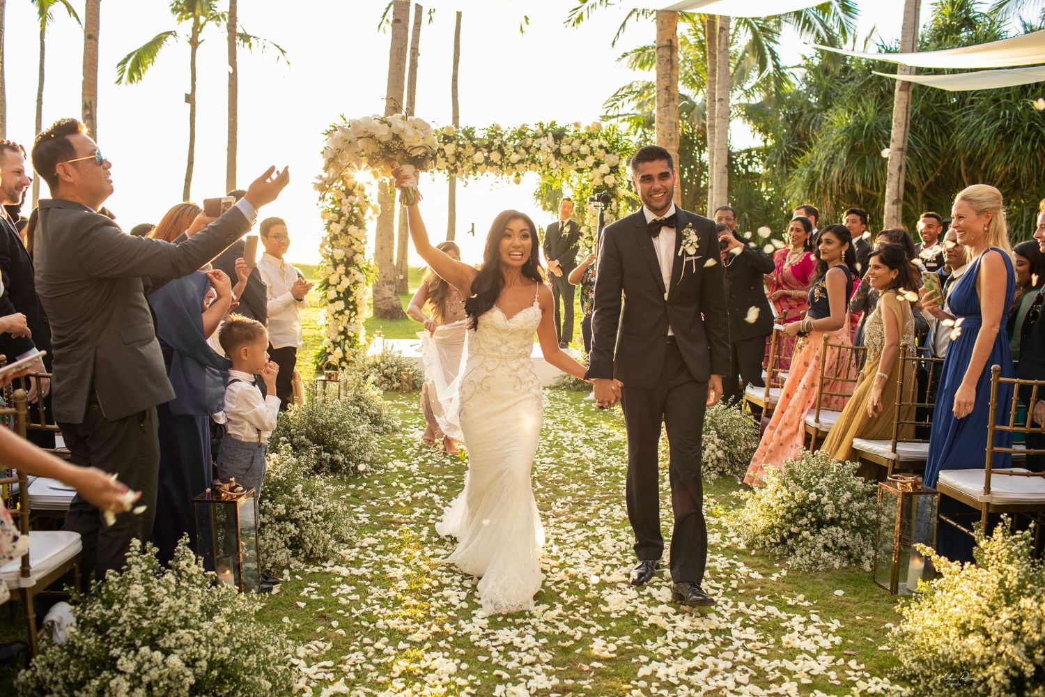 Newlyweds joyfully walking down a flower petal aisle as guests cheer at a tropical outdoor wedding.