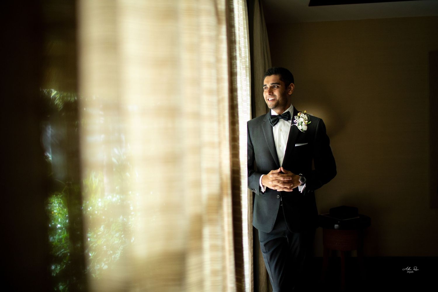 Dapper groom in black tuxedo with boutonniere gazes thoughtfully out window in softly lit room.