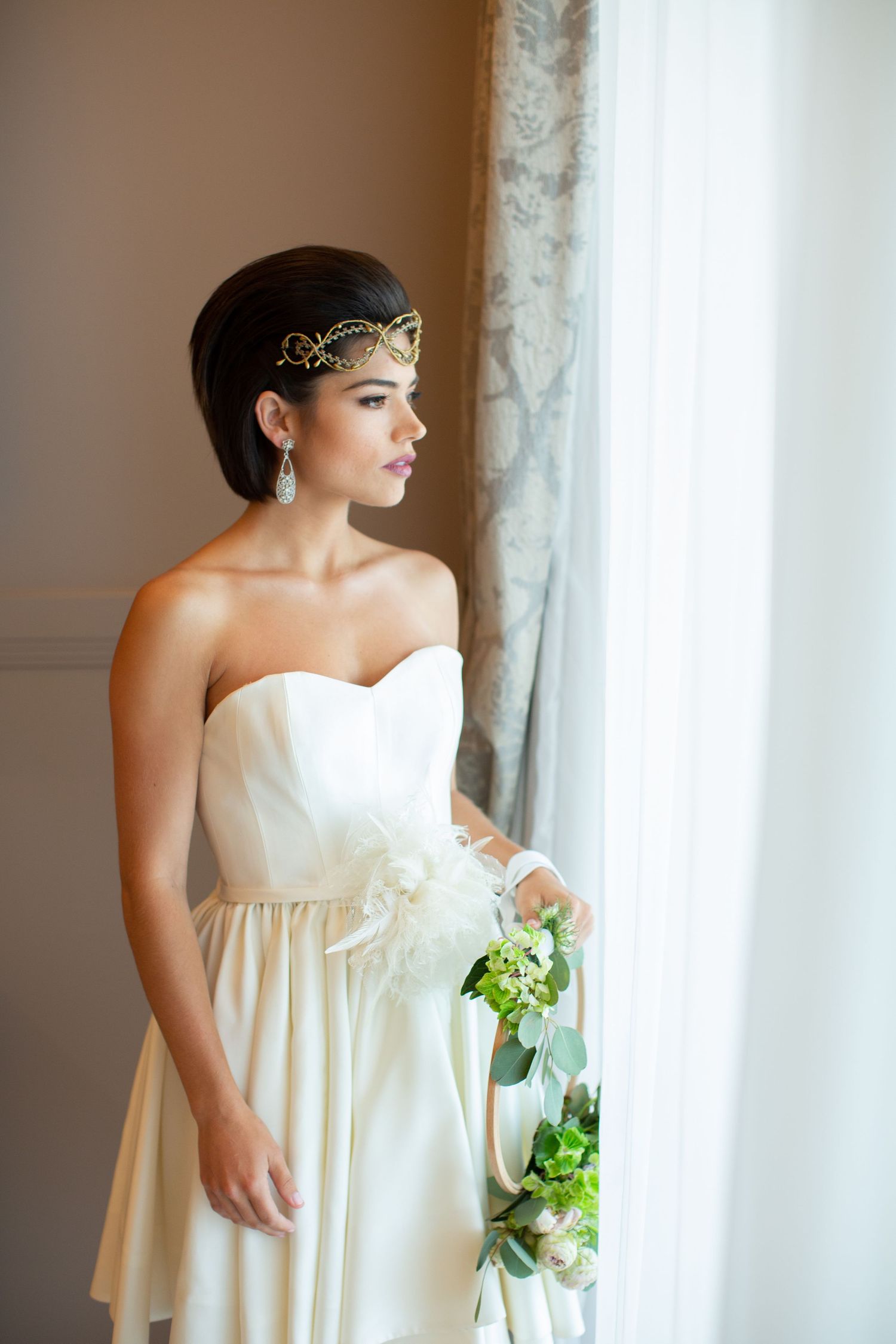 Sophisticated bride wearing a gold crown headpiece and strapless white gown holds a green bouquet by a window.