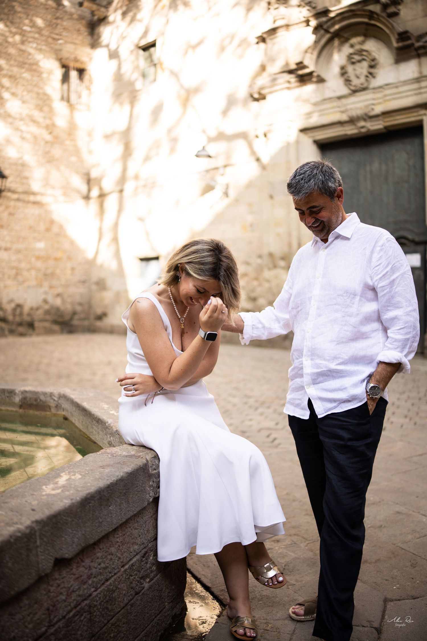 A couple shares a tender laughing moment beside a stone fountain in a sunlit historic Barcelona courtyard.
