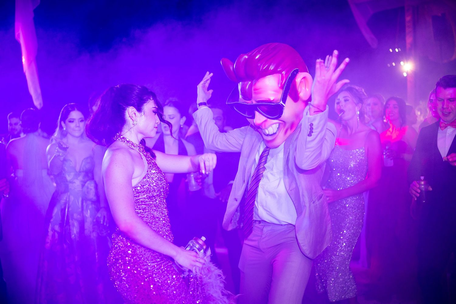 Energetic wedding guest in aviator goggles dancing under purple fog lights at a lively reception.