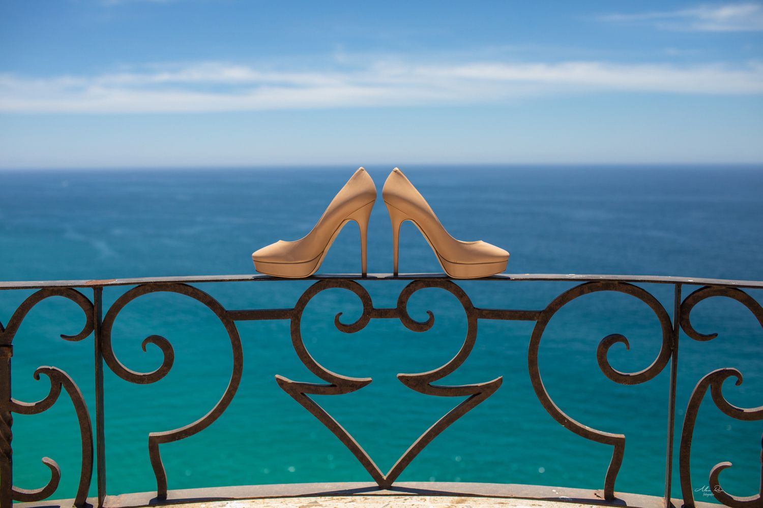 Nude high heel wedding shoes balanced on ornate iron balcony railing overlooking turquoise ocean.
