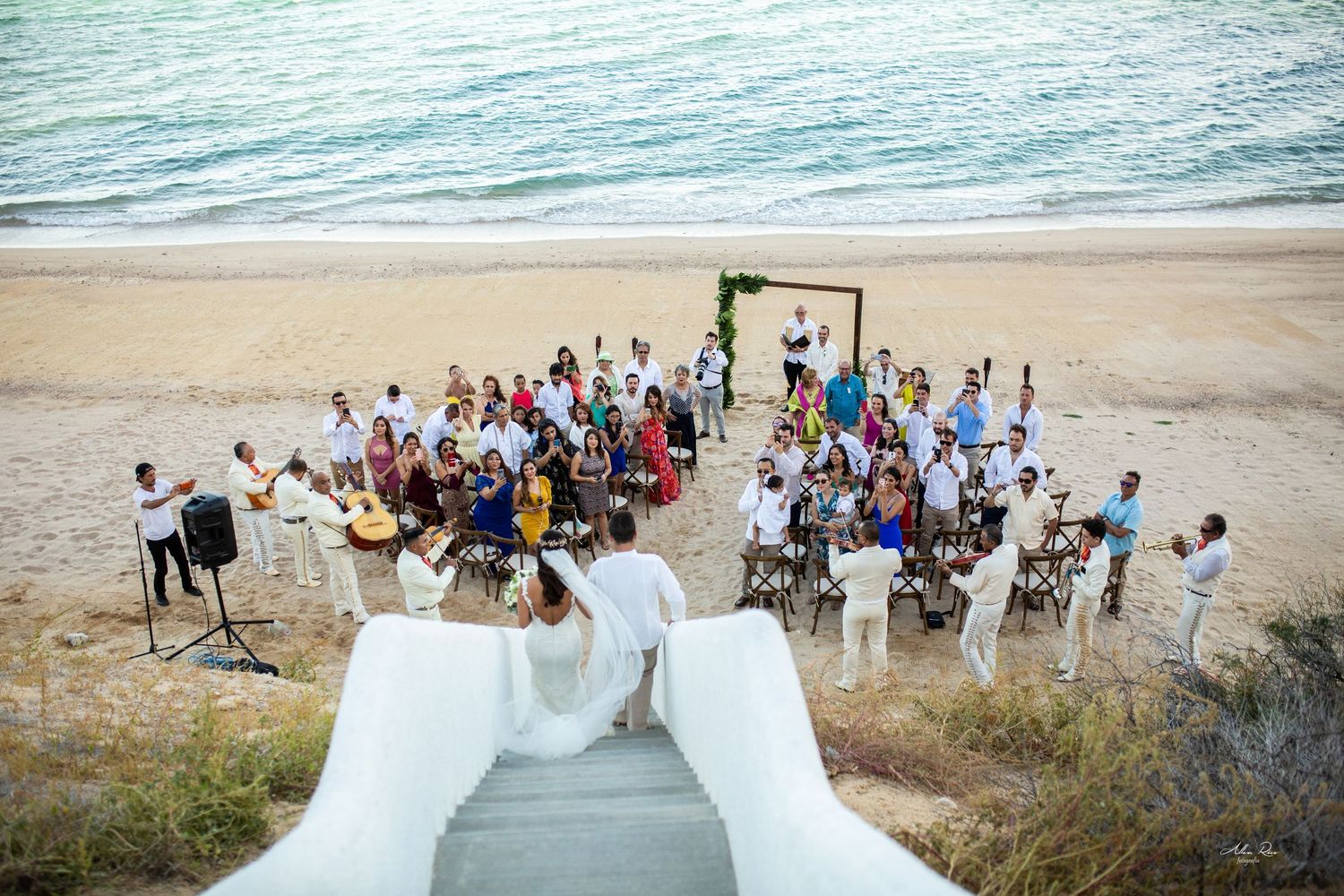 Aerial view of a beach wedding ceremony with guests gathered near the ocean shore on sandy beach.