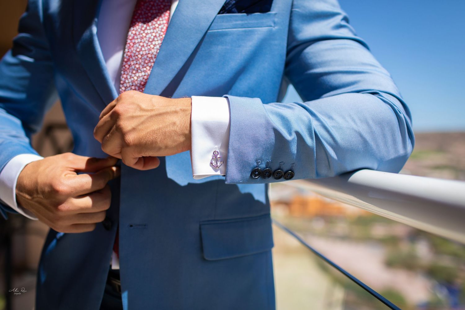 Close-up of groom in light blue suit adjusting cufflinks on outdoor balcony with city view.