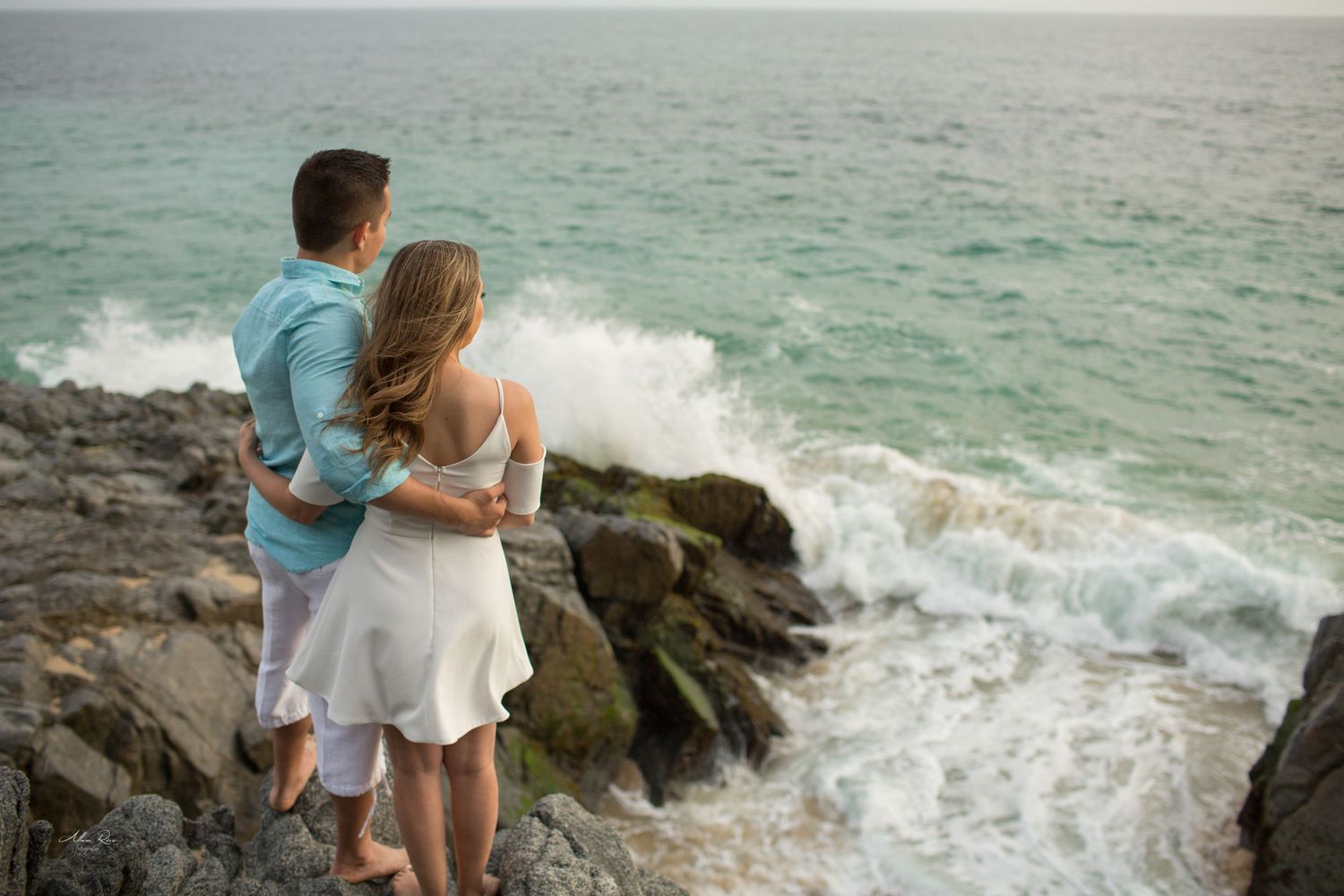 Couple embraces on rocky shoreline watching dramatic ocean waves crash against the coast.
