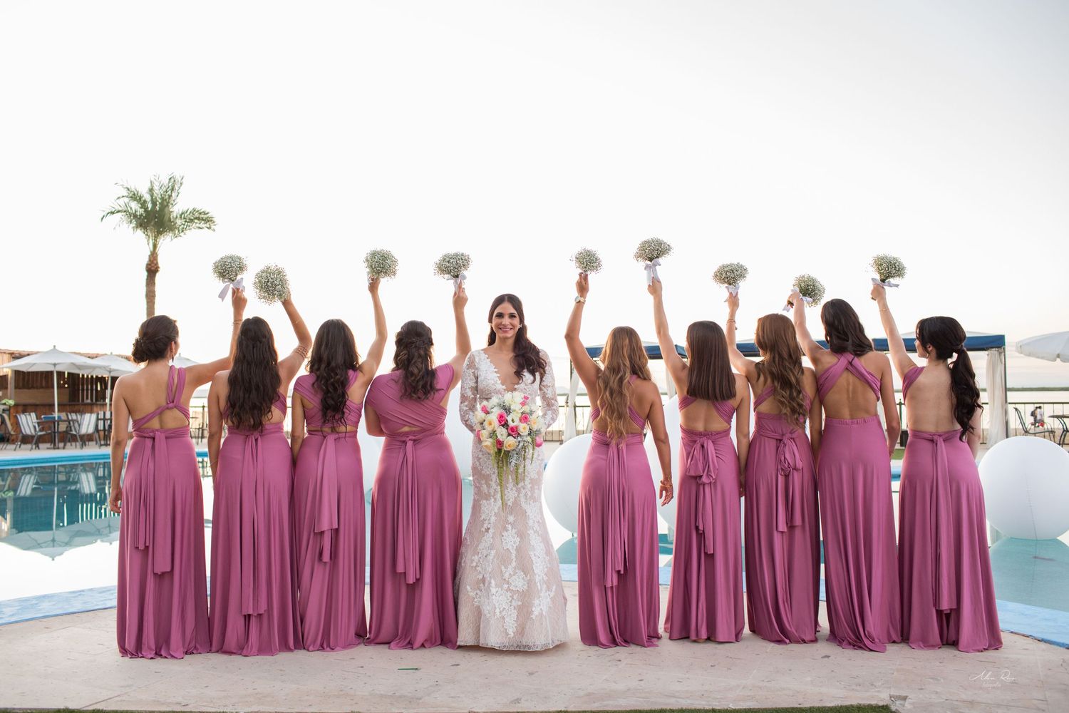 Bride in lace gown surrounded by bridesmaids in mauve dresses raising bouquets by resort pool.