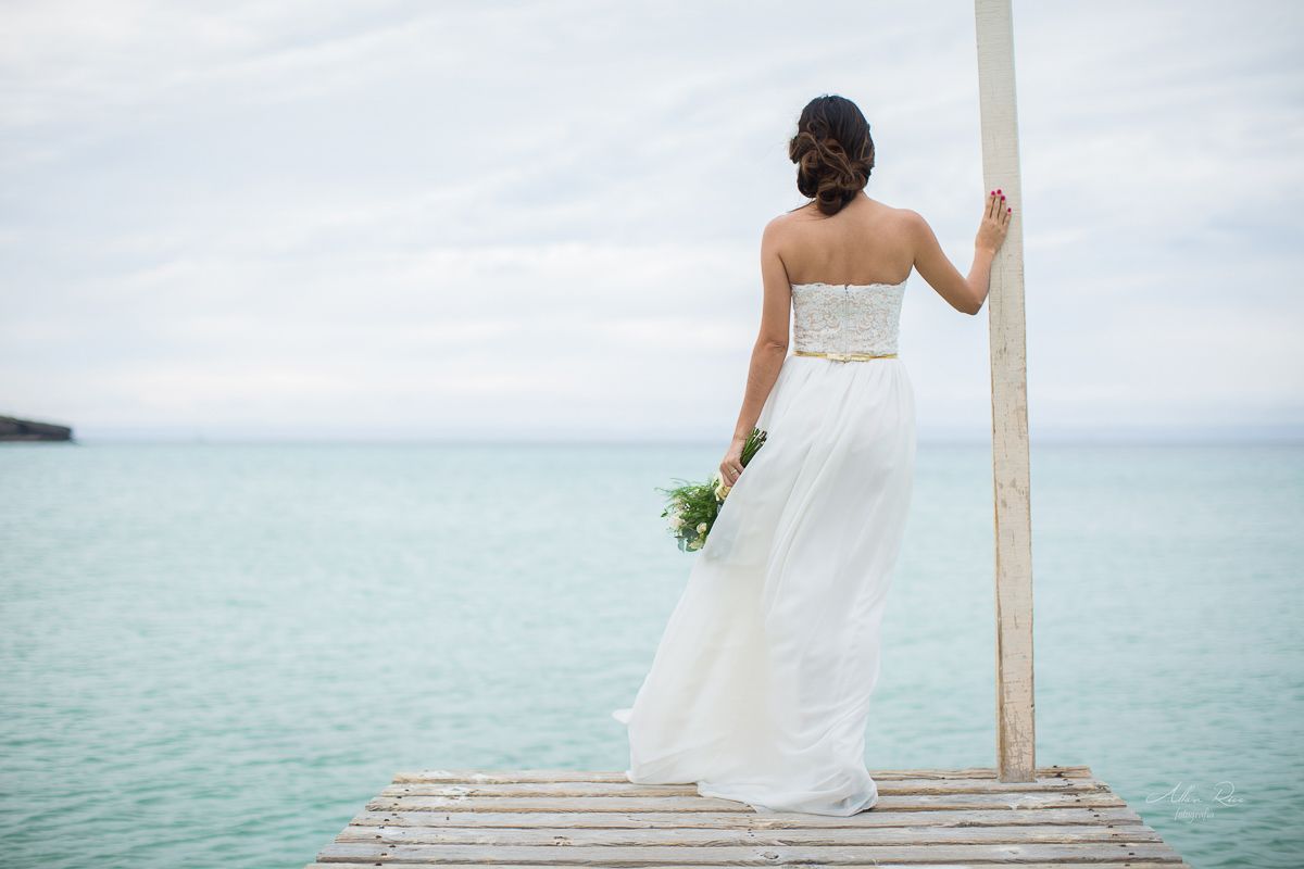 Bride in strapless white gown viewed from behind on wooden pier overlooking calm turquoise ocean.