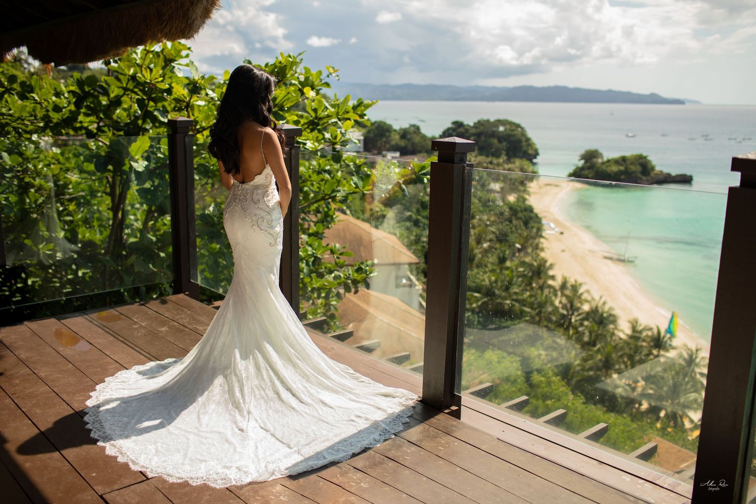 Bride in lace mermaid wedding dress poses on wooden deck with stunning tropical beach panorama behind her.