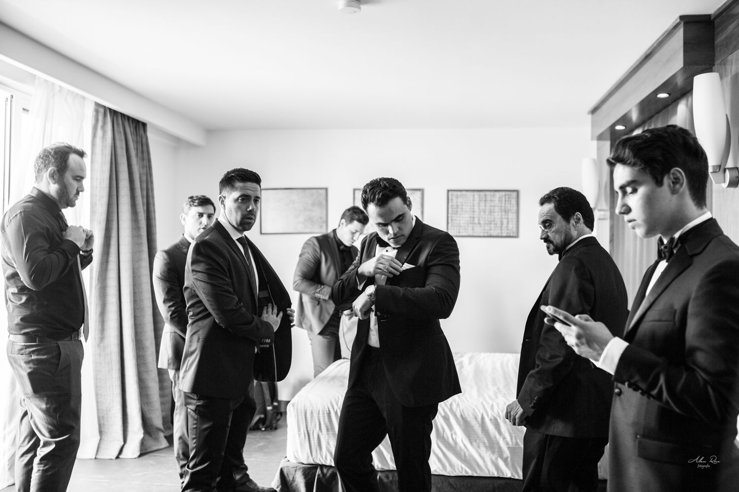Groomsmen in black tuxedos preparing together in hotel room before wedding ceremony.
