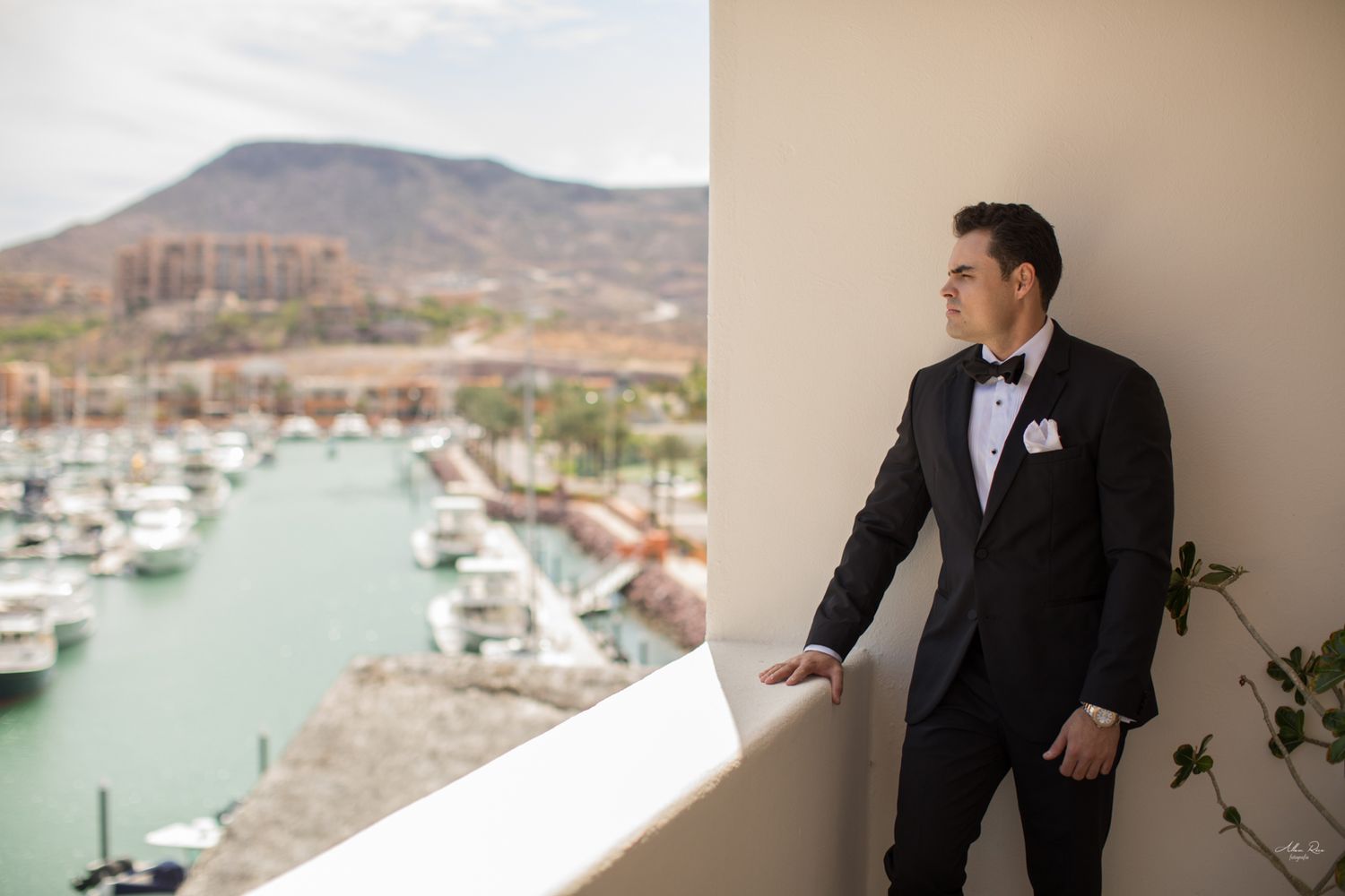 Groom in black tuxedo posing on balcony overlooking scenic marina and mountain landscape.