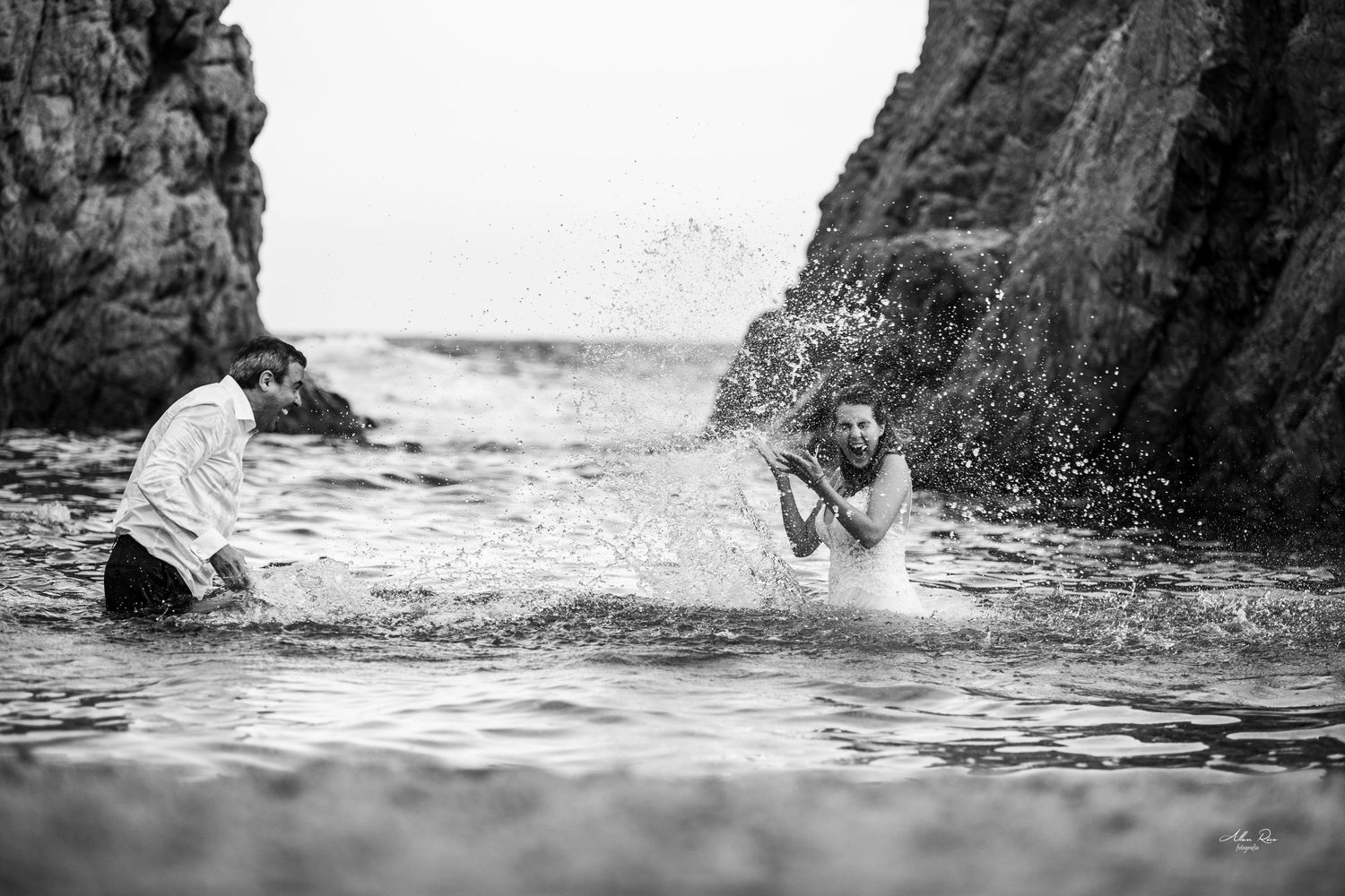 Bride and groom playfully splash in ocean waves between rocky cliffs in black and white.