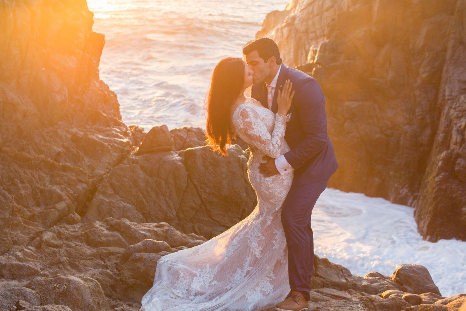 Wedding couple embracing passionately on ocean rocks during breathtaking golden hour sunset.