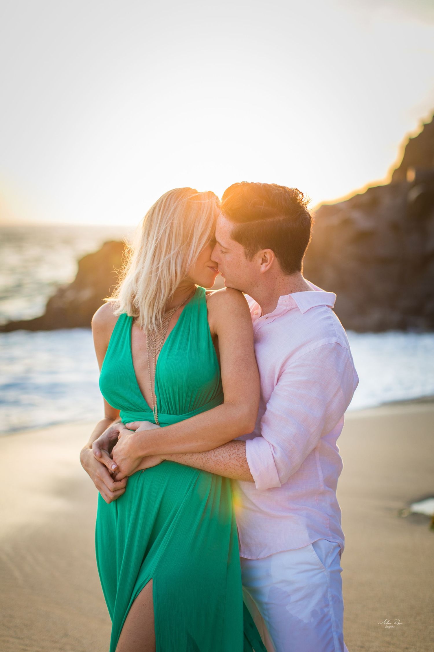 Couple embracing and almost kissing on a beach at sunset, woman in green dress, man in pink shirt.