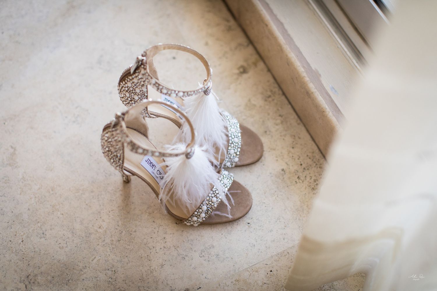 Ornate crystal and feather bridal heels displayed on a marble windowsill beside a wedding dress.