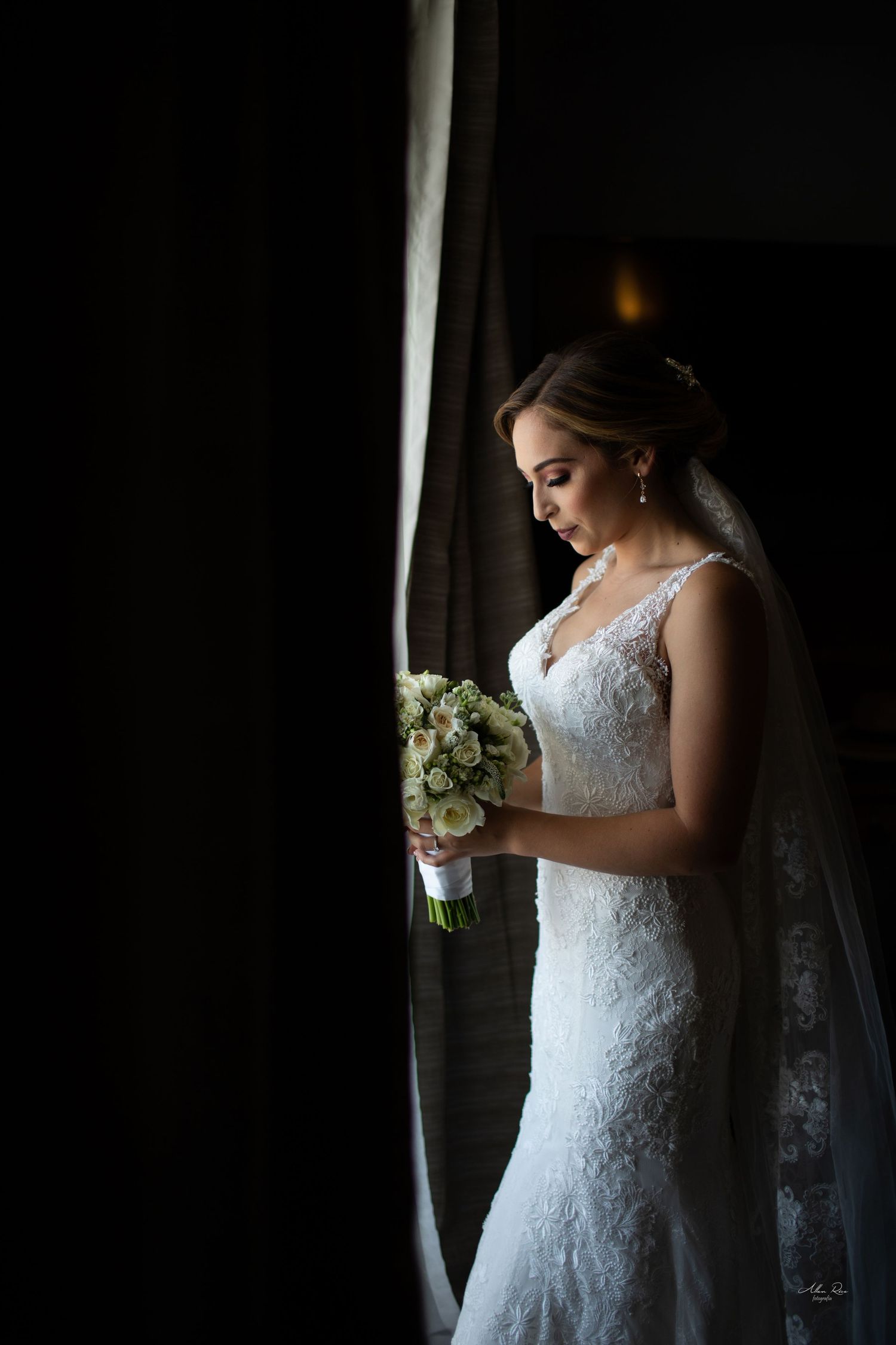 Beautiful bride in lace wedding gown holding a white bouquet while standing by a dark curtained window.