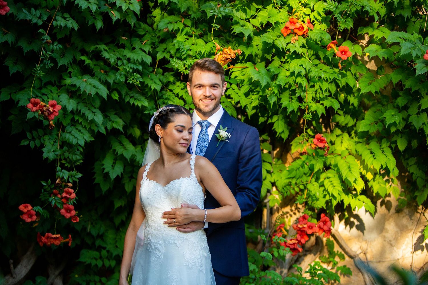 Smiling bride and groom embracing in front of lush green ivy with red flowers.
