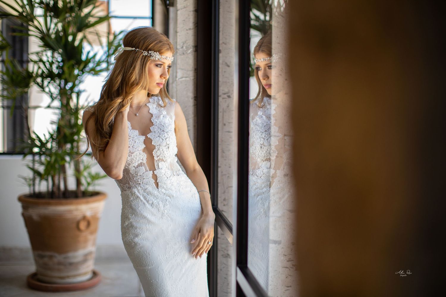 Elegant bride in fitted lace wedding gown standing by glass door with reflection visible.