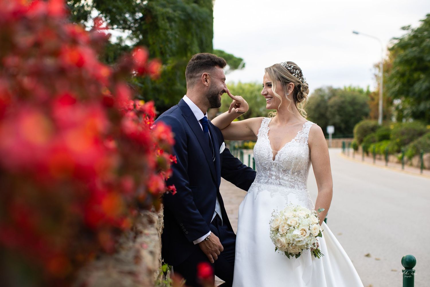 Smiling bride and groom pose near vibrant red flowers with bouquet of white roses outdoors.
