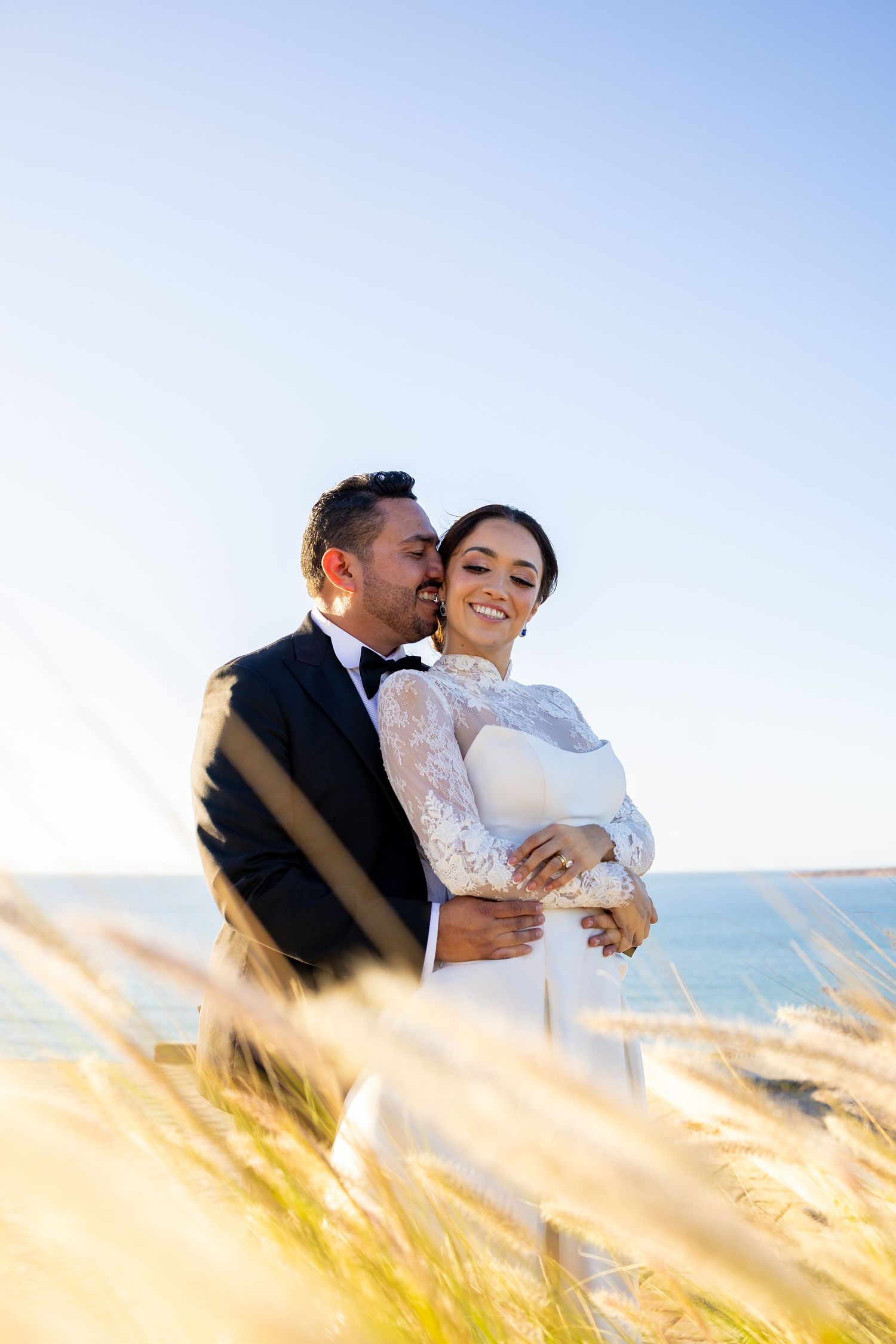 Groom embracing smiling bride among golden coastal grass with ocean view in background.