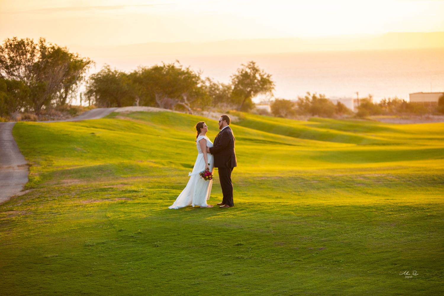 Bride and groom stand facing each other on a scenic golf course at golden sunset with ocean views.