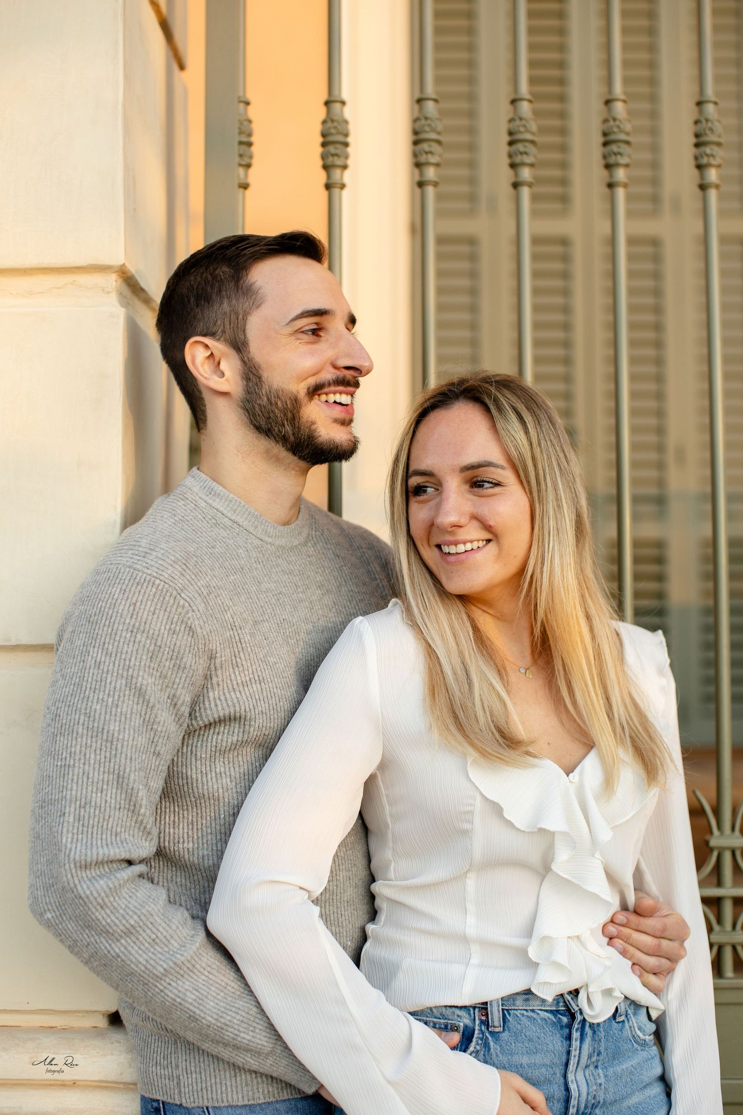 Smiling couple embracing outdoors near a vintage building, man in grey sweater, woman in white blouse.