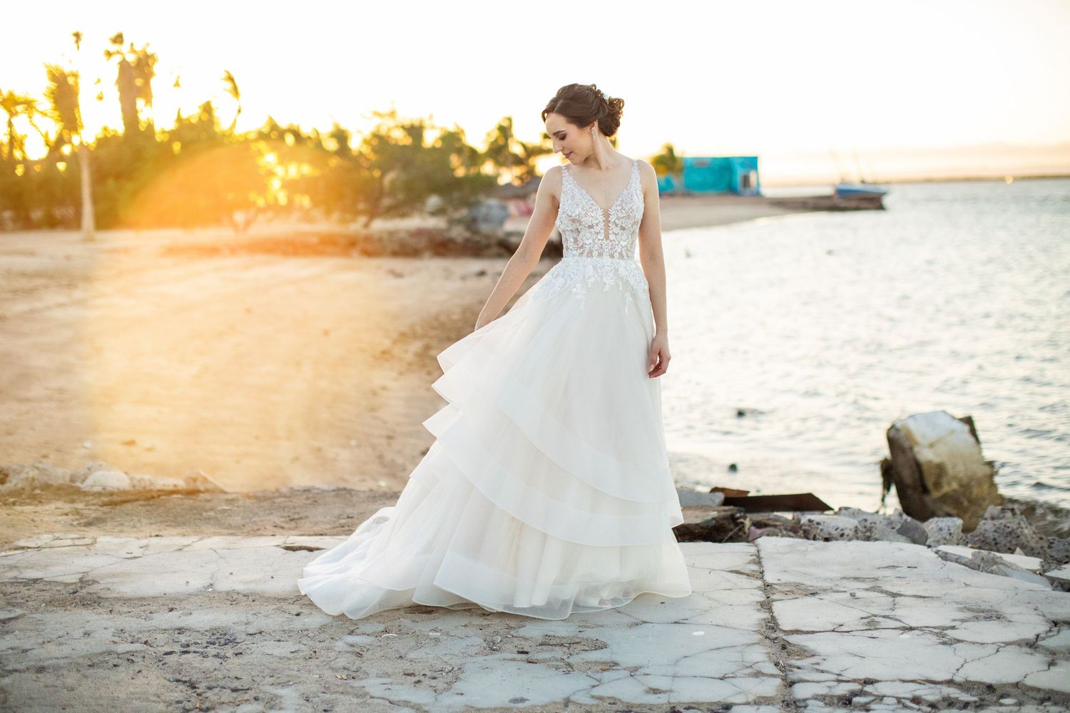 Bride in tiered white wedding gown walking on rocky shore bathed in golden sunset light.