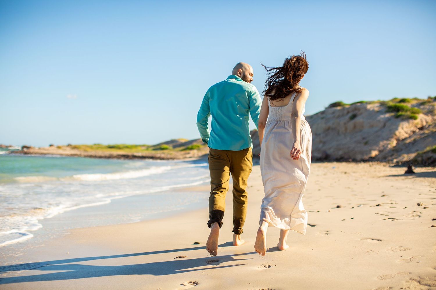 Couple running barefoot along a sunny sandy beach holding hands.