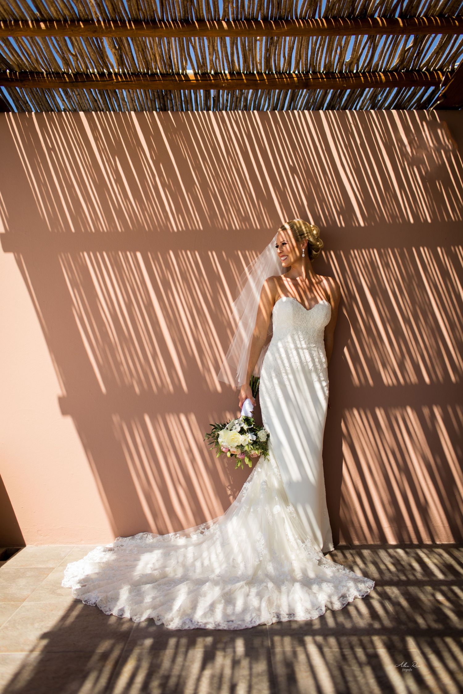 Bride in lace wedding gown posing against terracotta wall with dramatic shadow patterns.