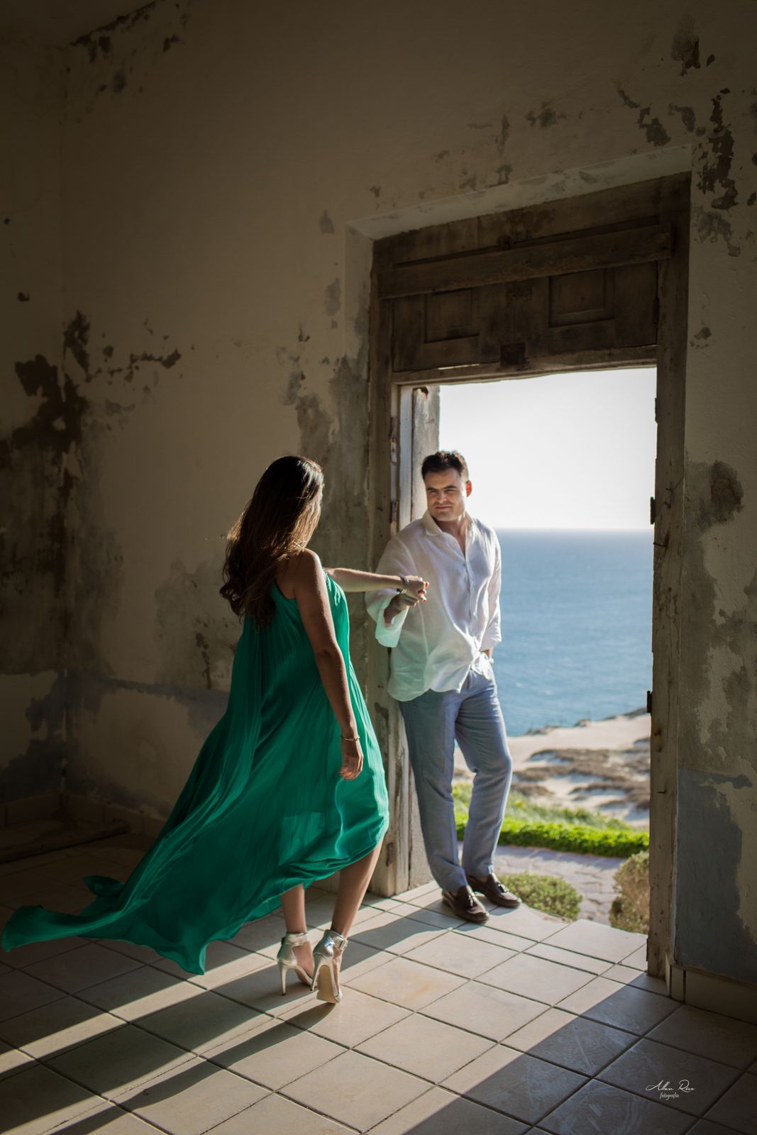 Couple dancing in doorway of rustic abandoned building with ocean view, woman in elegant green gown.