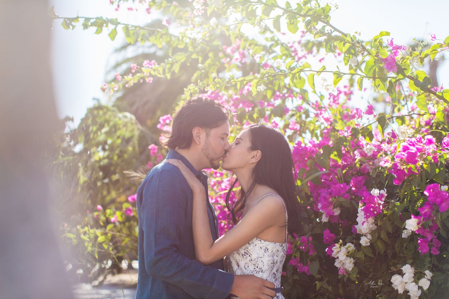 Couple shares a tender kiss surrounded by vibrant pink bougainvillea flowers in sunlight.