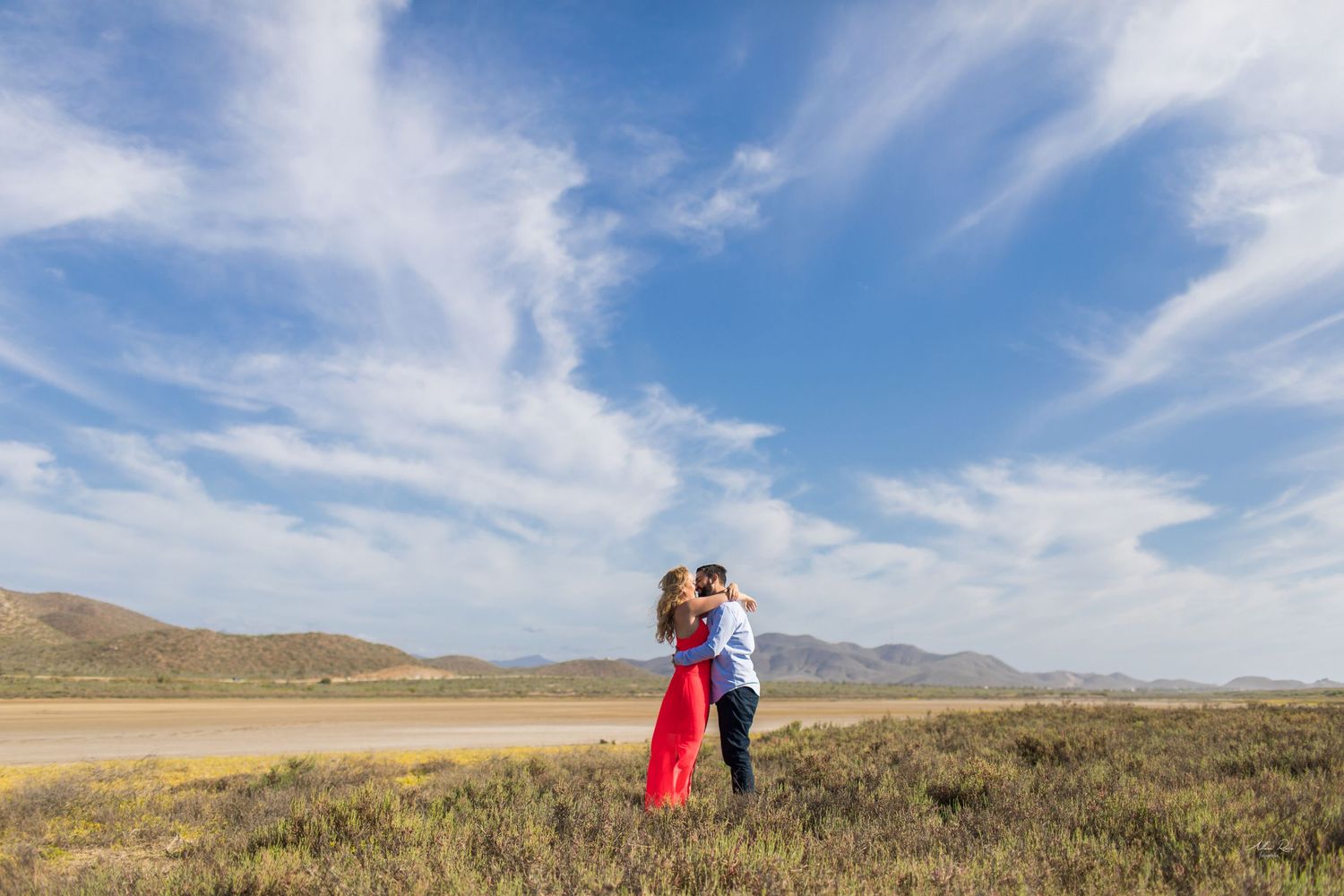 Romantic couple kissing in open desert field under stunning blue sky with wispy clouds.