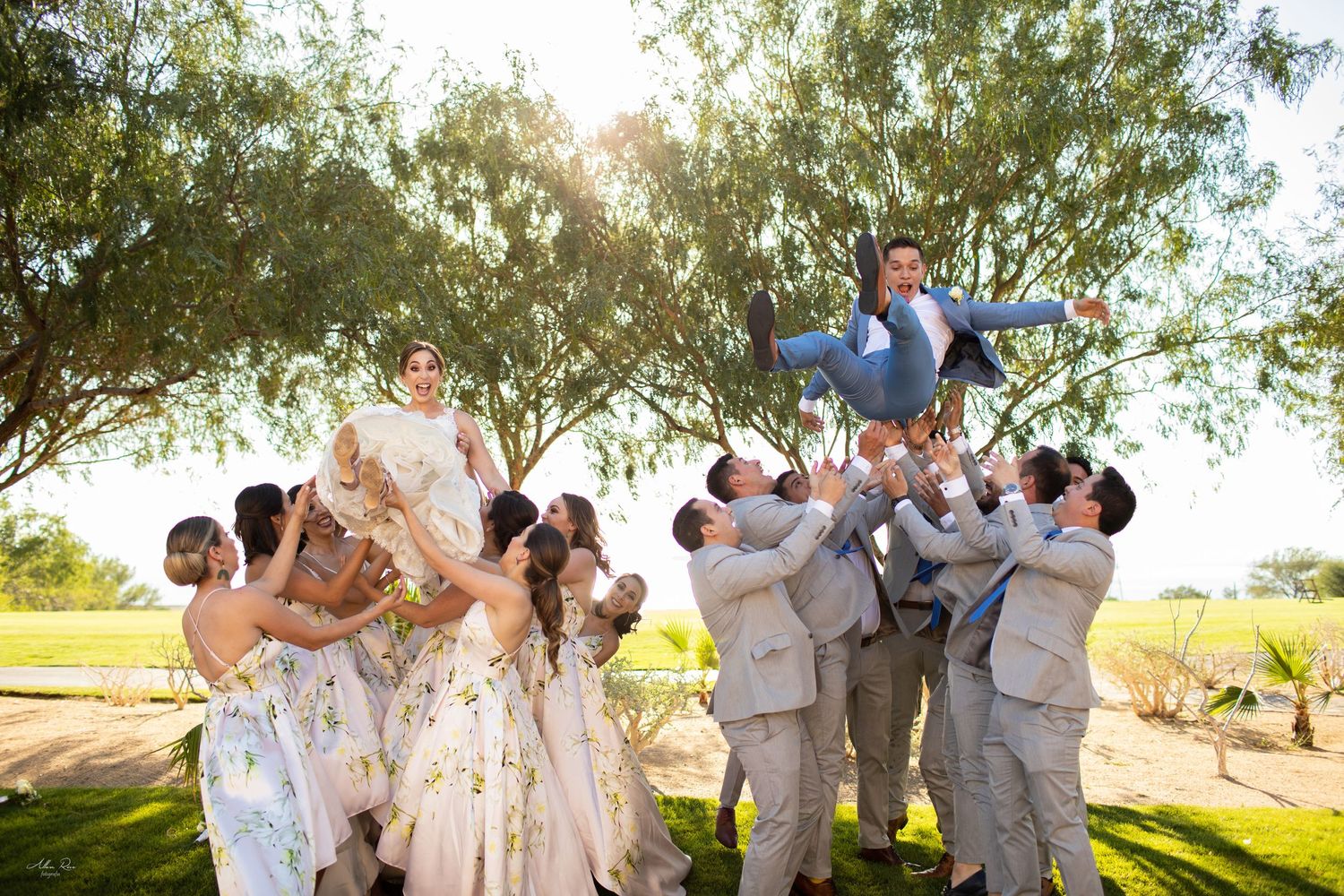 Wedding party joyfully tosses bride and groom into the air together during a sunny outdoor celebration.