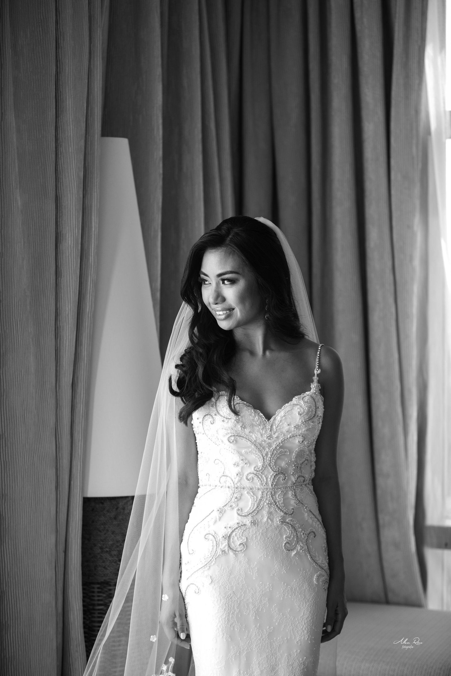 Black and white portrait of a beautiful bride in a lace fitted gown and veil standing by a window.