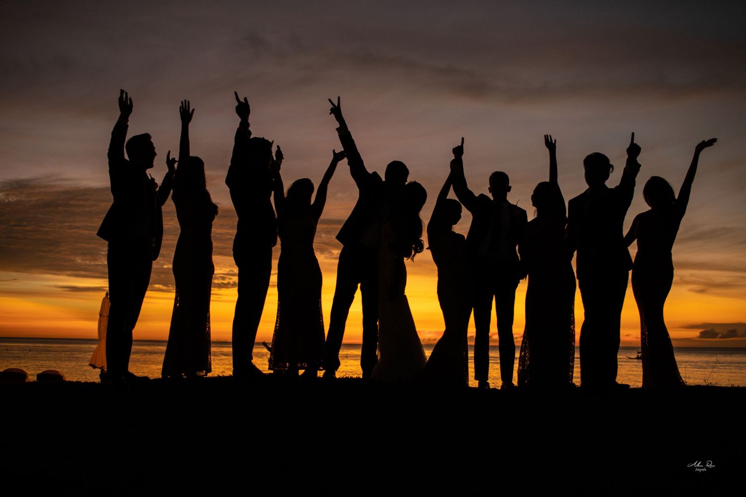 Silhouettes of a wedding party striking fun poses against a vibrant orange and gold sunset sky.