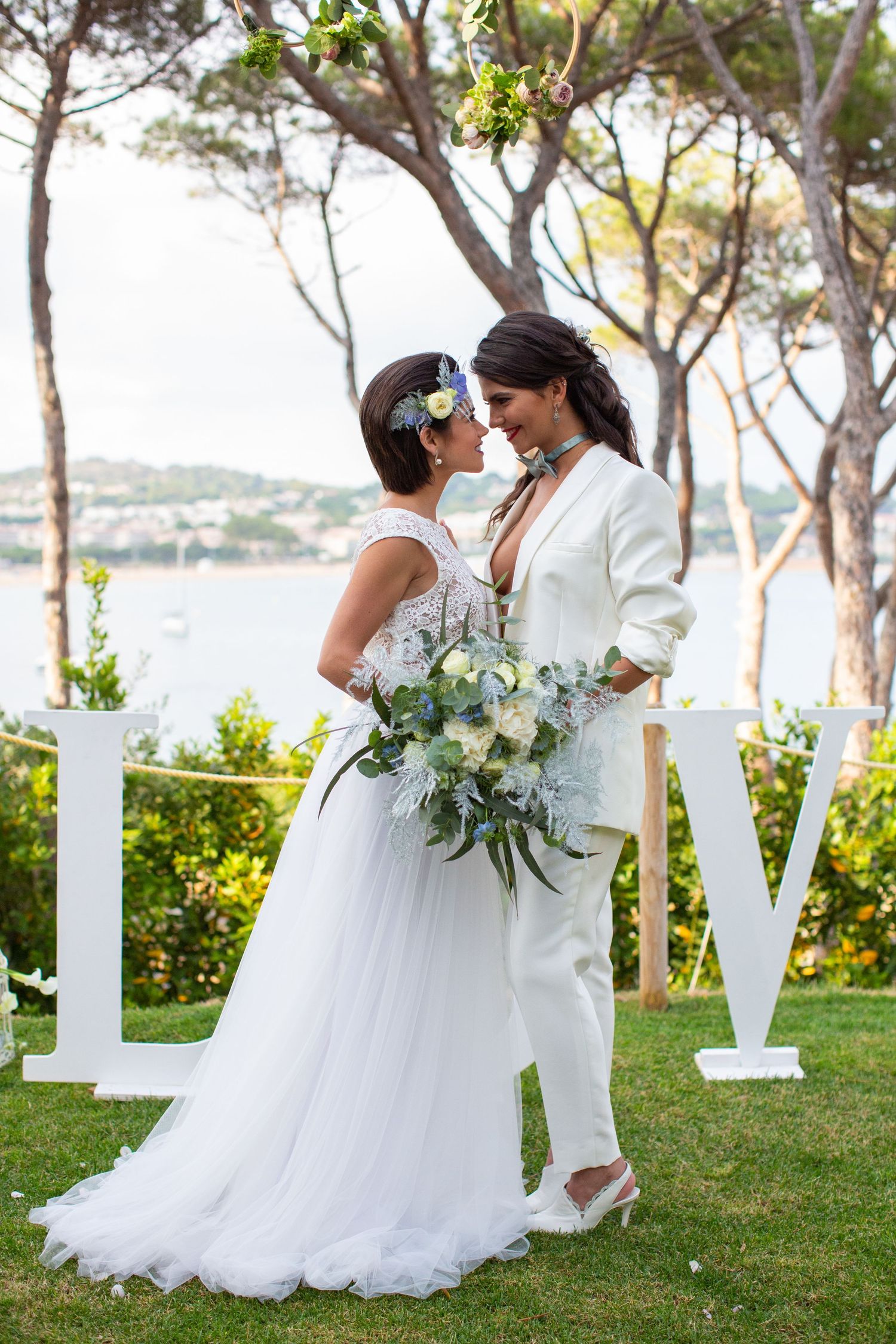 Two brides in white wedding attire share a romantic moment outdoors near LOVE letters with ocean views.