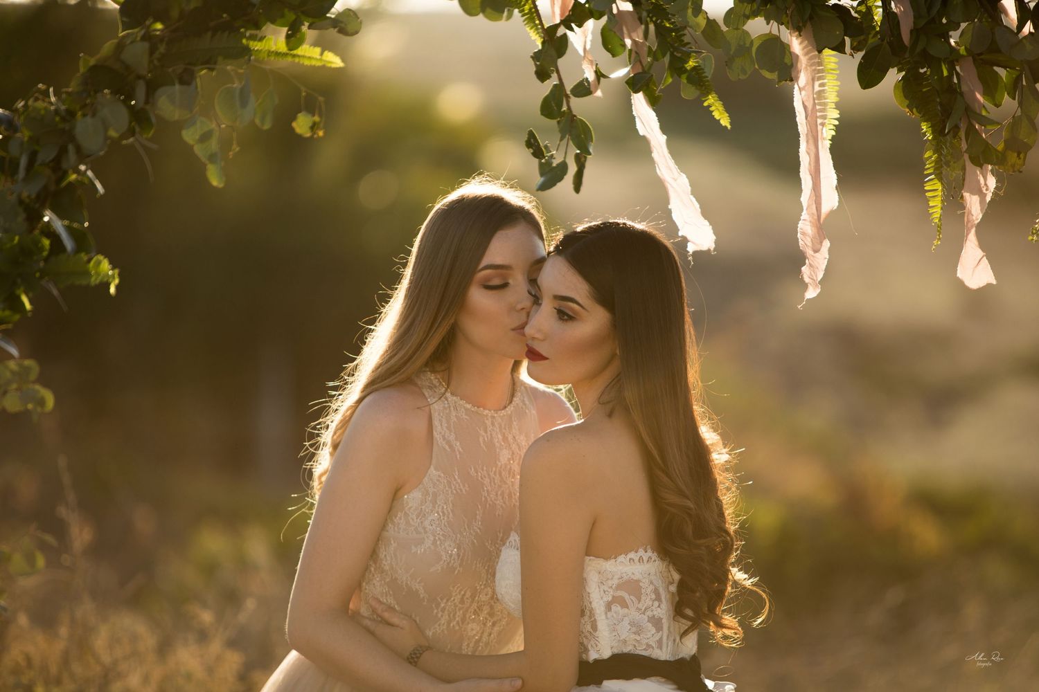 Two brides sharing a gentle kiss under sun-drenched hanging branches at golden hour.