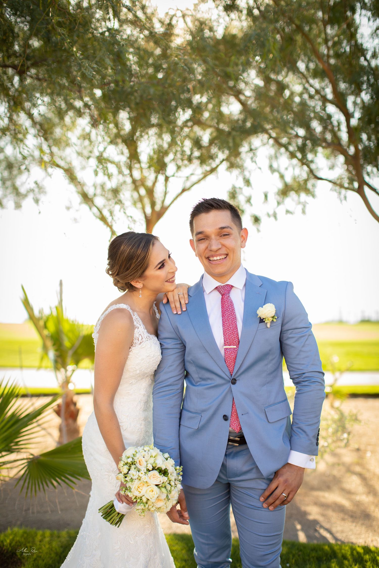 Smiling groom in blue suit with pink tie poses with bride holding white bouquet under a palm tree.