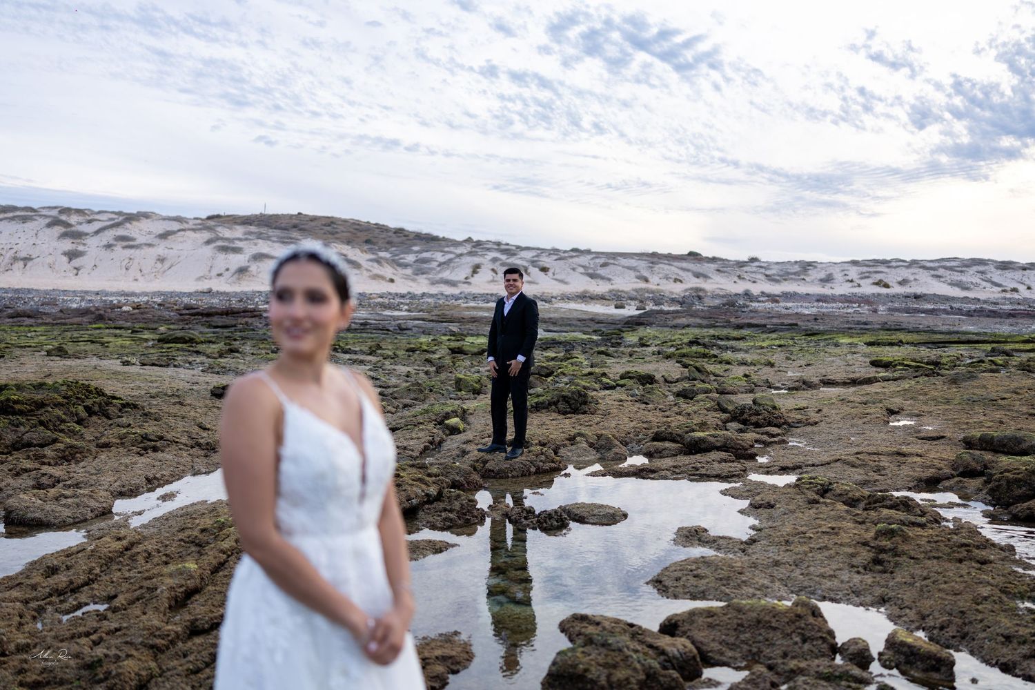 Bride and groom posing on rocky coastal landscape with sand dunes at dusk.