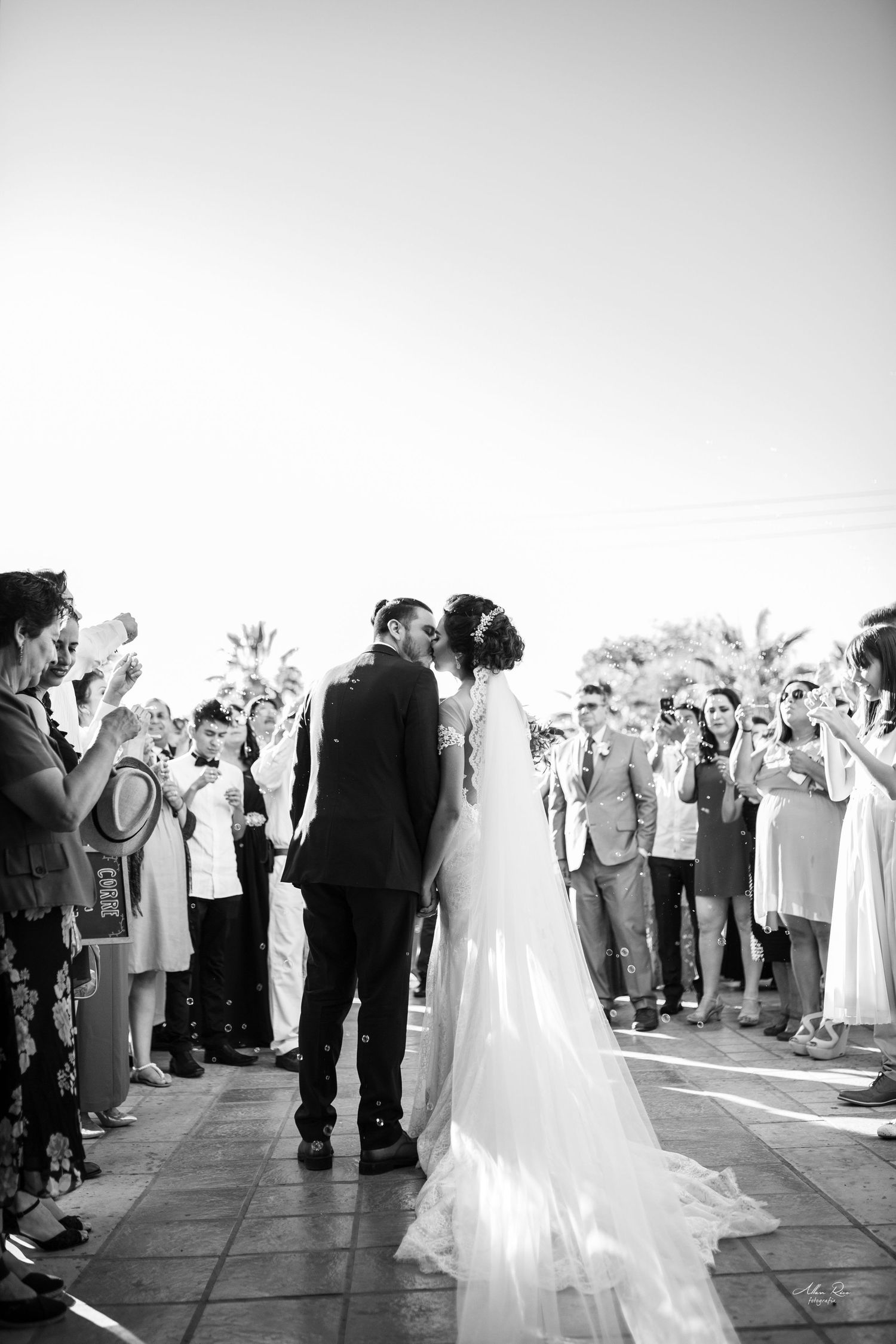 Newlyweds share their first kiss as a married couple while guests celebrate outdoors in black and white photo.