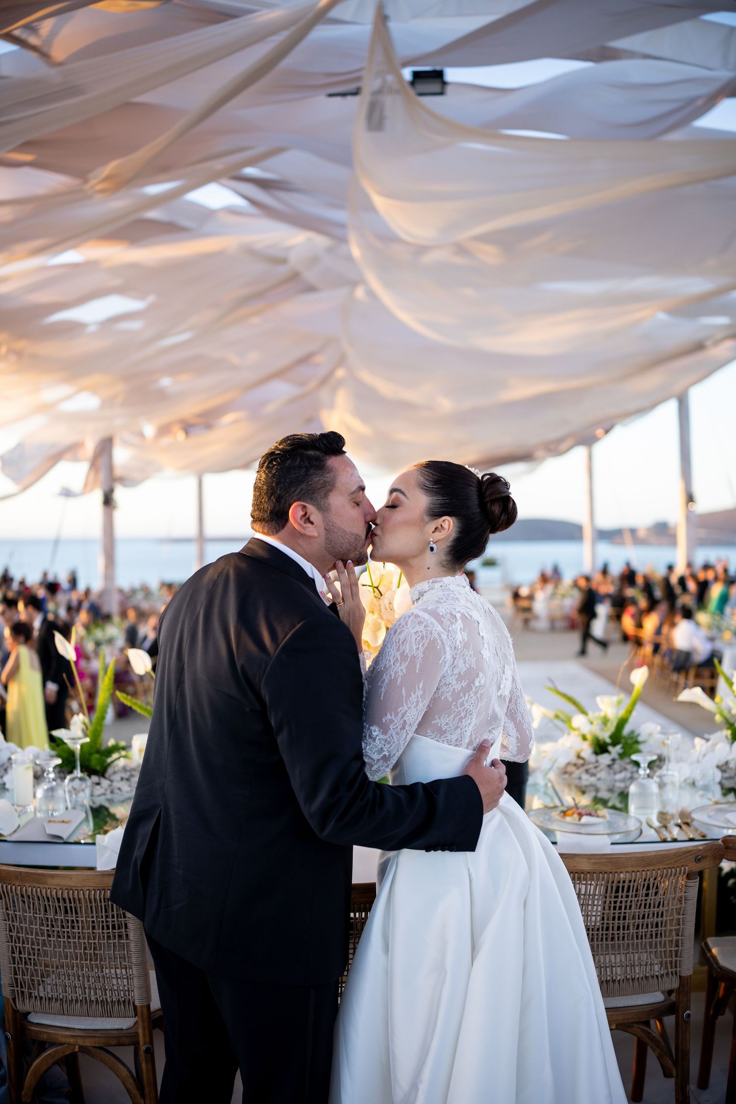 Newlyweds share romantic kiss under elegant white draped canopy at beachside wedding reception.