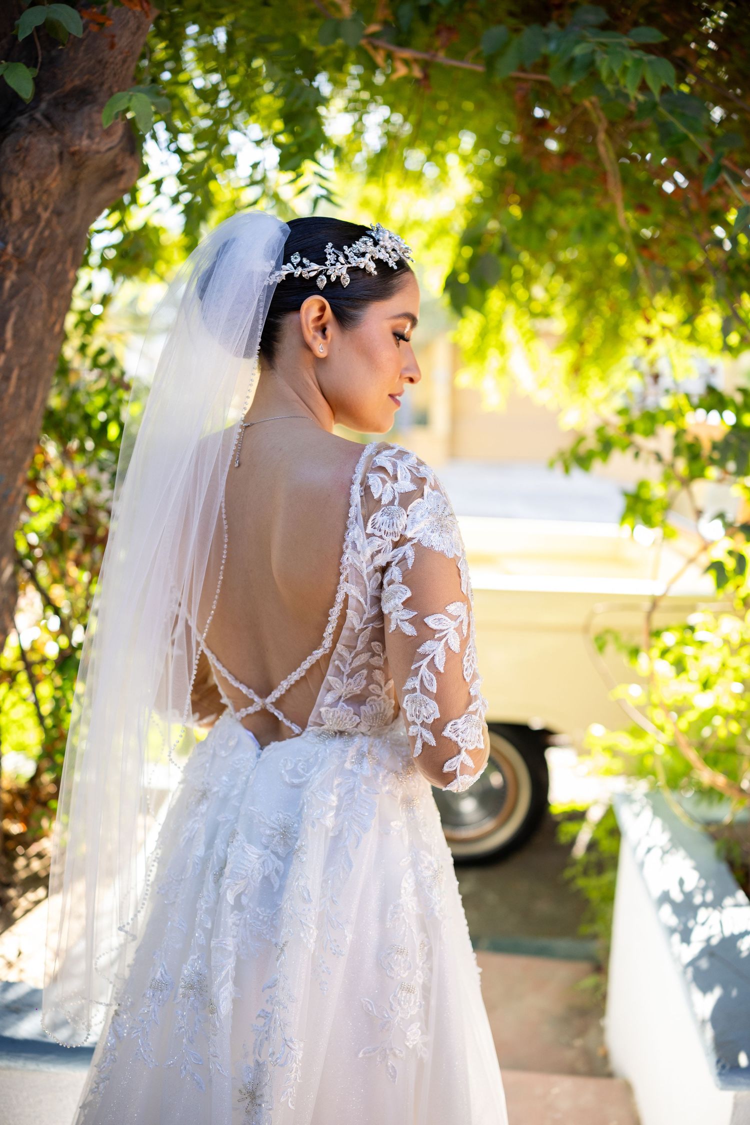 Bride in backless lace wedding dress with veil and tiara standing under green trees outdoors.