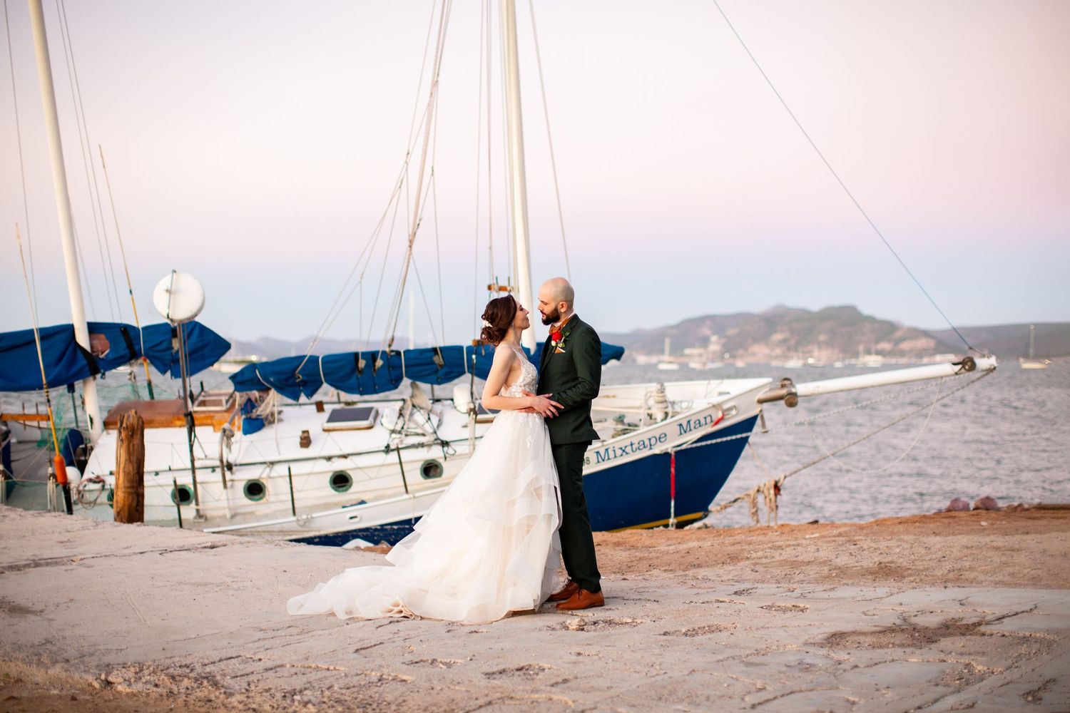 Bride and groom share a tender kiss near a sailboat docked on a calm waterfront at dusk.